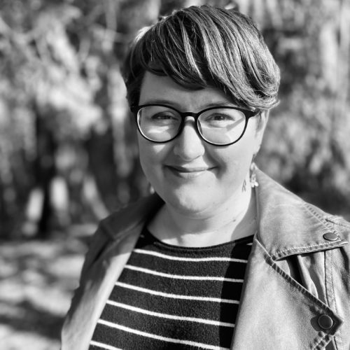 black and white photo of Karen Olson, a woman with short hair wearing a striped shirt and jacket standing in the woods